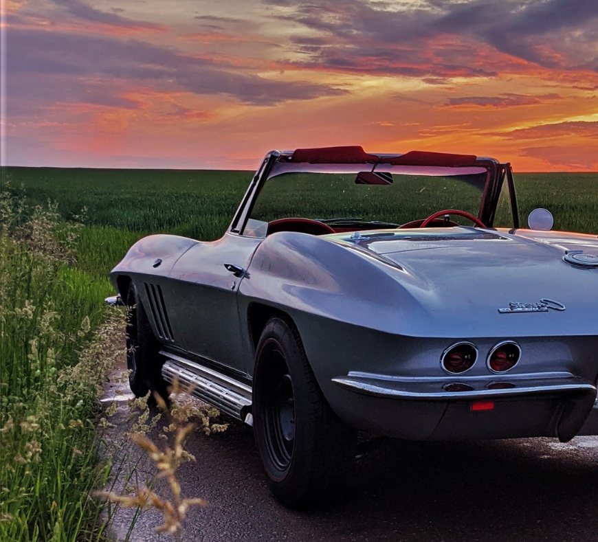 A classic car cruising down a scenic road, surrounded by lush grassy fields, embodying the timeless elegance of vintage automotive design.