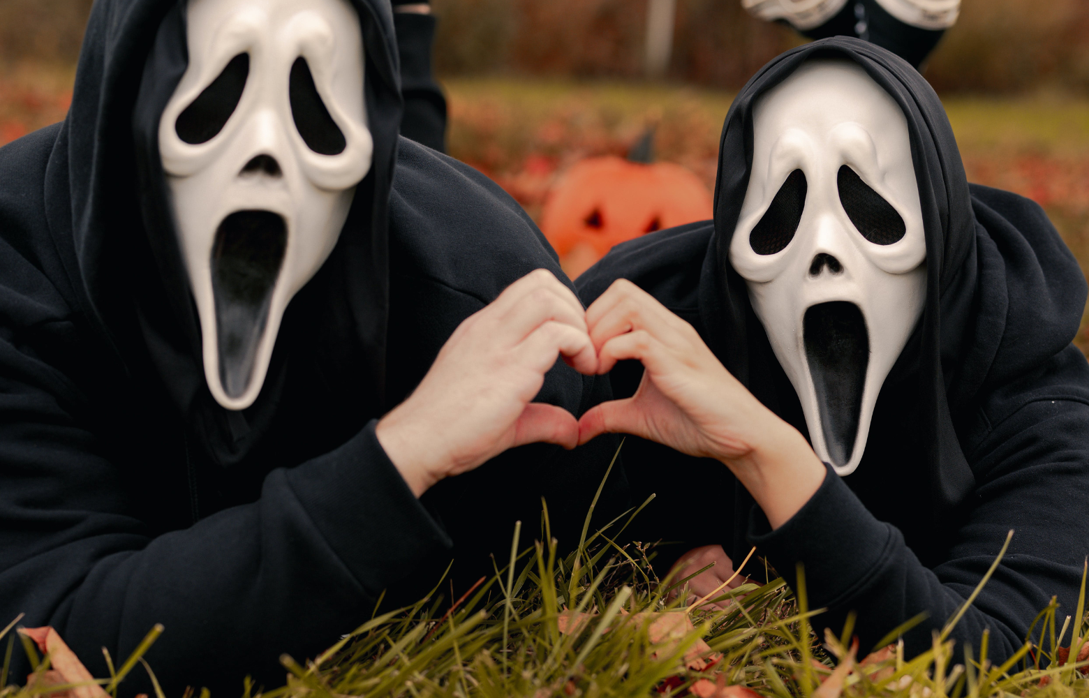 Two people wearing ghost masks lie side by side on the ground, their hands touching to form the shape of a heart, blending eerie and sentimental elements.